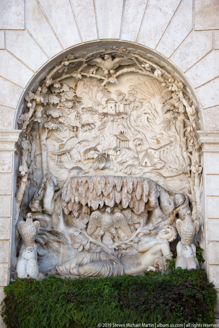 Large stone relief in an arched niche with mountains, buildings, vegetation, and a reclining figure above greenery.