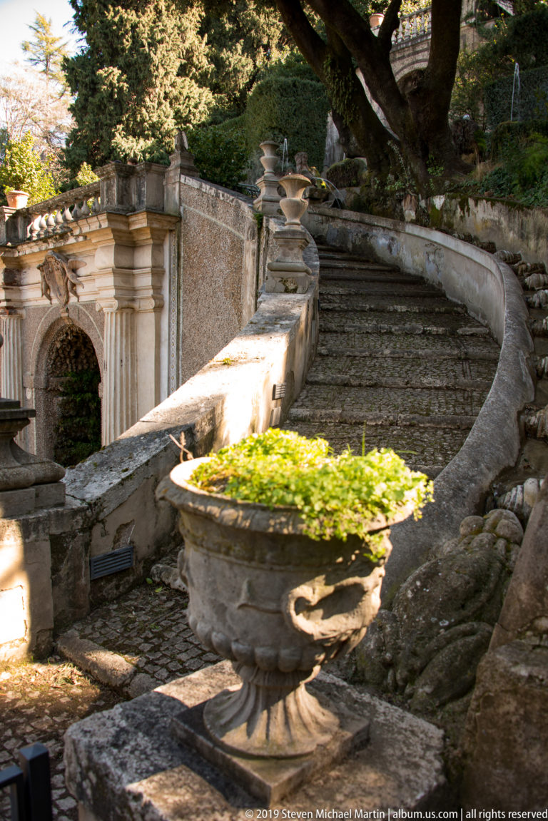Large planted stone urn before a curved weathered staircase and low parapet in a shaded garden.