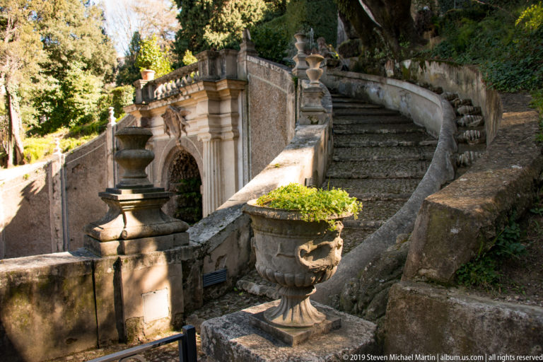 Curved stone staircase beside balustrades, urns, and an arched garden structure in dappled light.