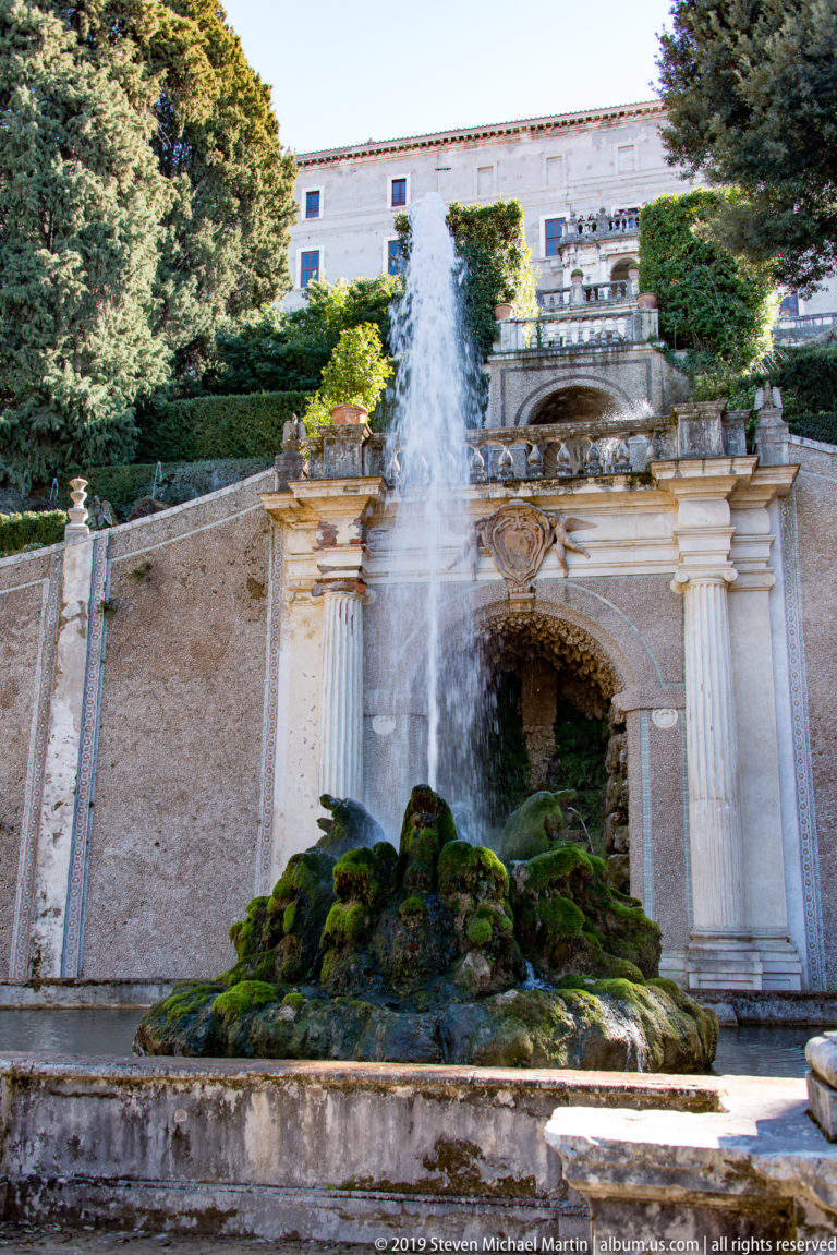 Vertical water jet rises from mossy rockwork before a wall with pilasters and an arched recess.
