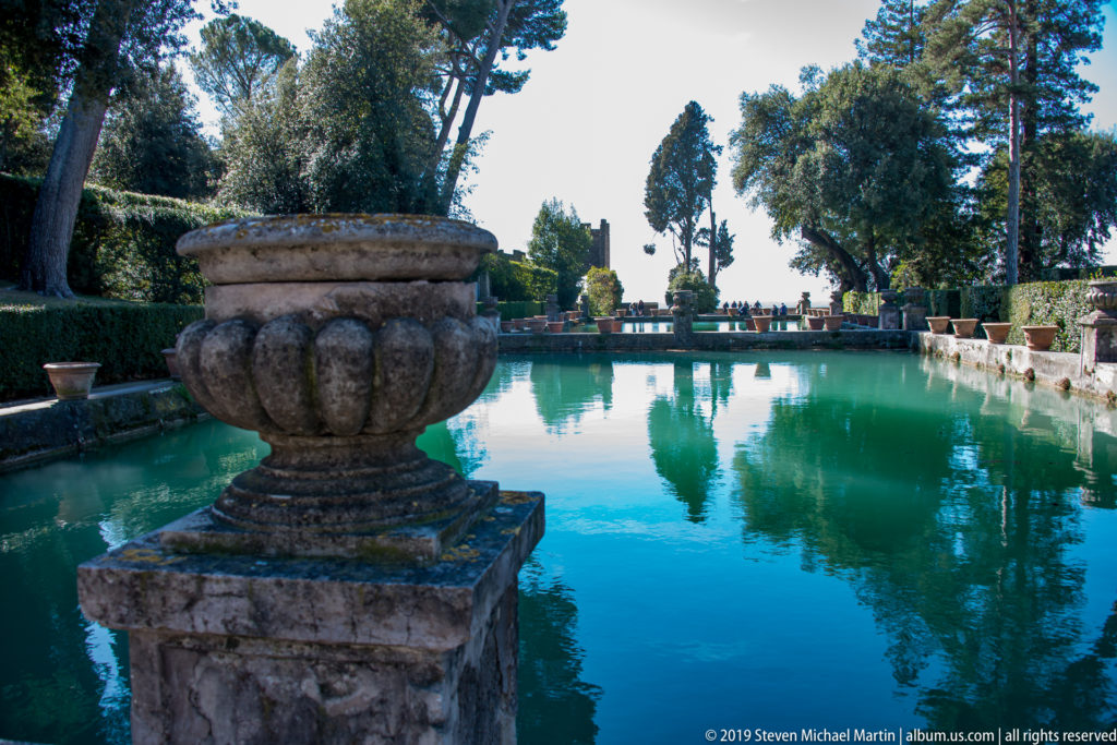 Large carved stone urn beside a long turquoise rectangular pool lined with trees and clipped greenery.