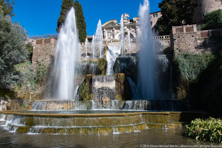 Twin vertical jets flank a broad tiered cascade beneath an ornate pale architectural fountain front.
