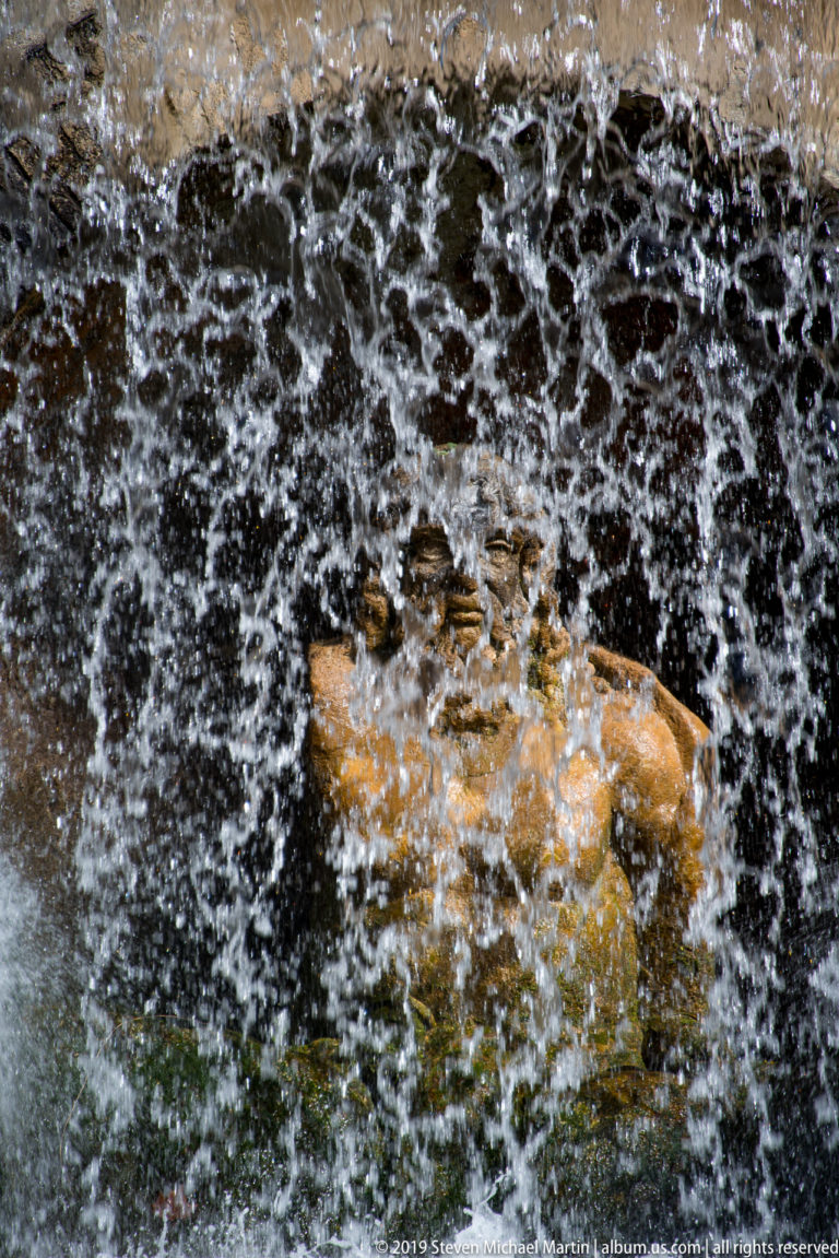 Heavy fountain water splashing over dark rock and a partly visible reclining stone figure.