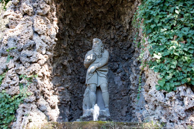Small stone figure holding an object in a rough grotto niche, with ivy at right and a water jet below.