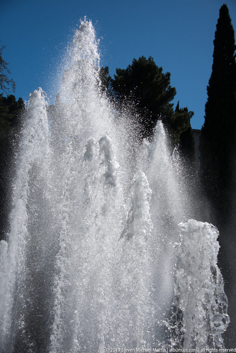 Dense overlapping fountain plumes and airborne droplets against dark trees and blue sky.