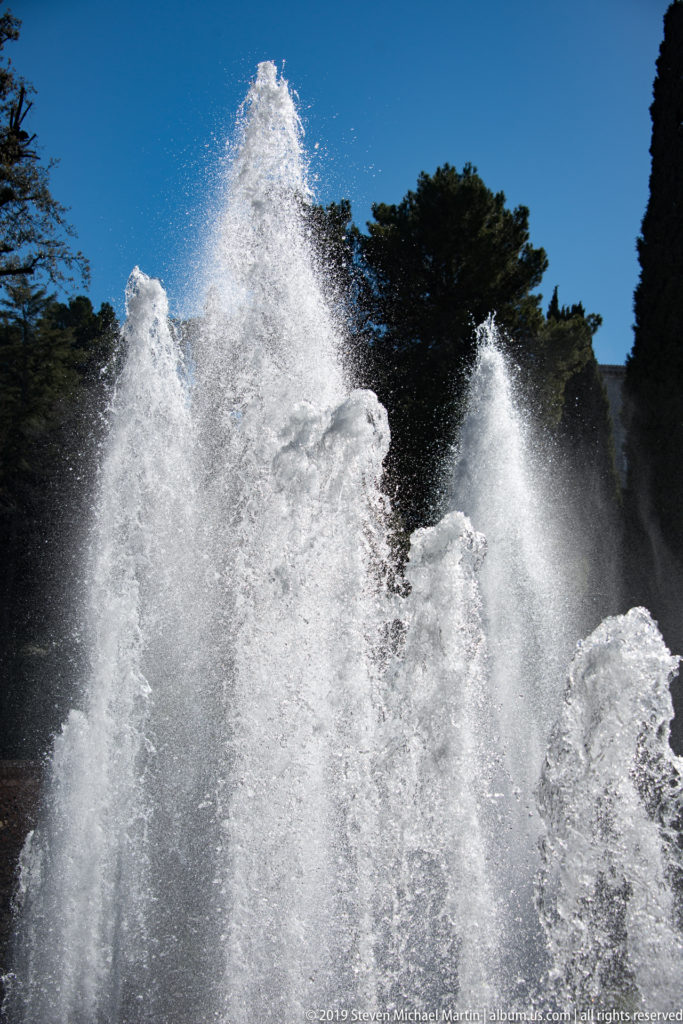 Cluster of powerful white fountain jets rising against a deep blue sky and dark trees.