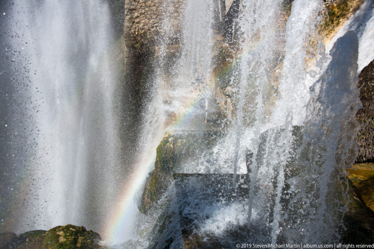Small rainbow visible through dense white fountain spray falling across dark rock and masonry.