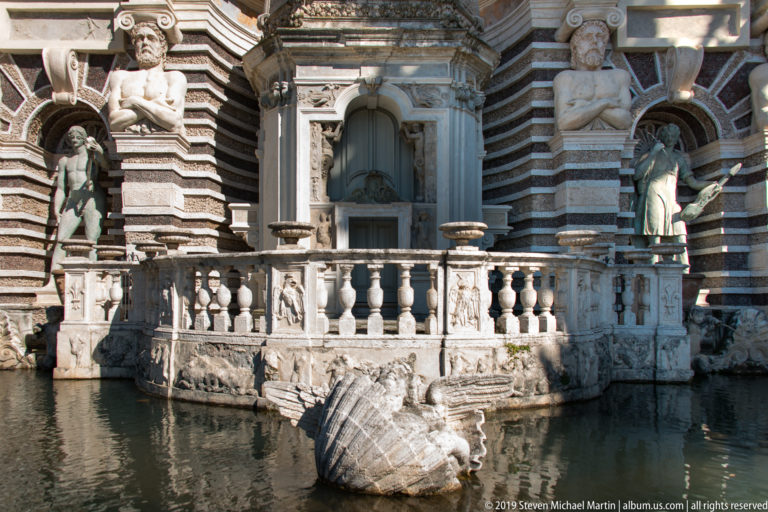 Balustraded fountain platform above a pool with large ribbed prow-like sculpture, striped masonry, and statues behind.