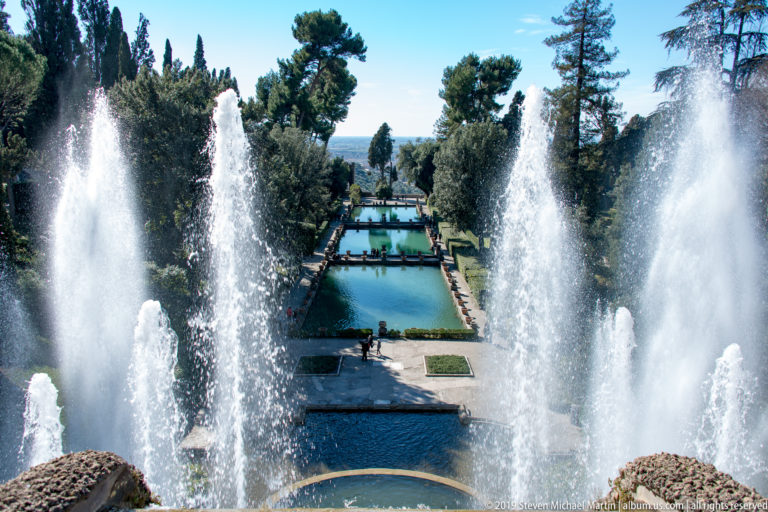 Tall fountain jets frame a descending central view of rectangular pools, paths, and trees in a formal garden.