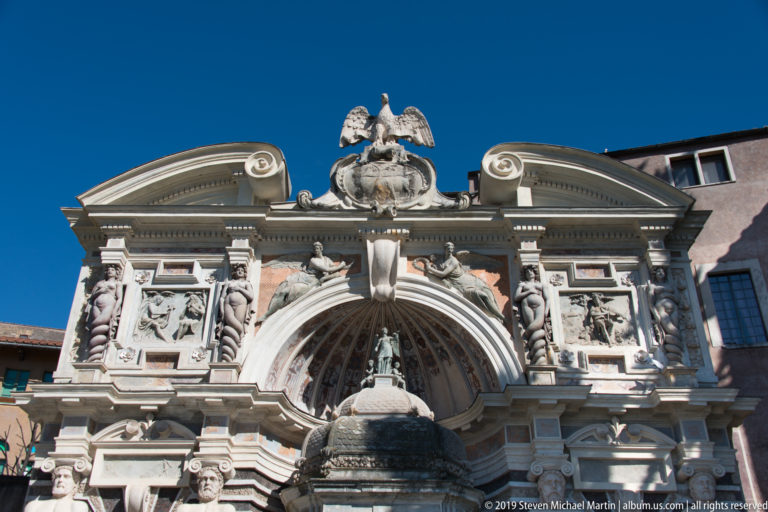 Upper portion of ornate fountain façade with shell recess, eagle crest, relief panels, statues, and curved pediments.
