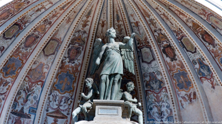Standing classical statue on a pedestal beneath a richly painted ribbed shell vault with smaller figures below.