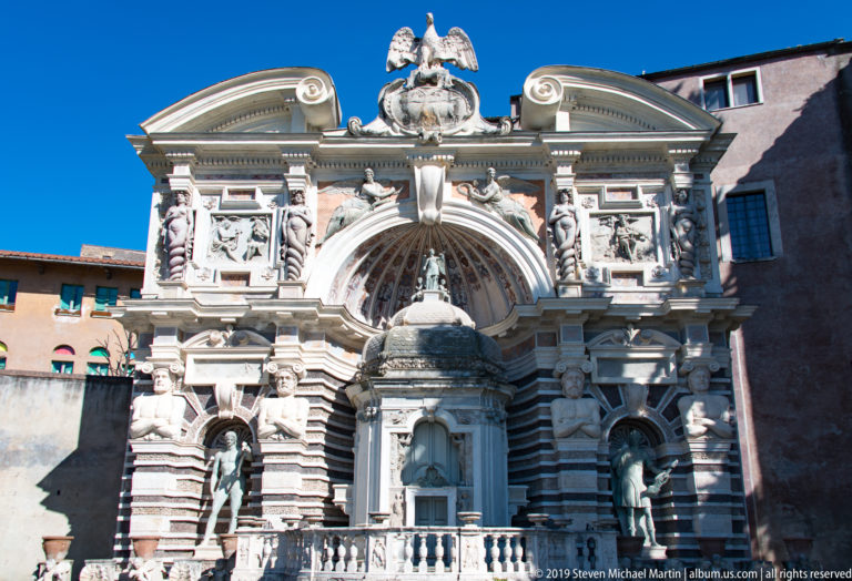 Ornate fountain façade with striped masonry, statues, relief panels, central shell recess, eagle crest, and basin.