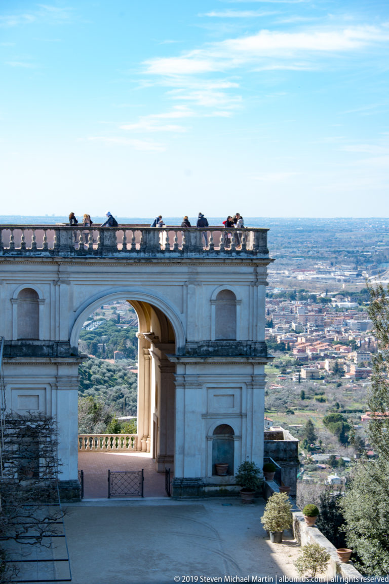 Pale stone terrace arch with visitors on the upper balustrade overlooking the expansive plain below Tivoli.