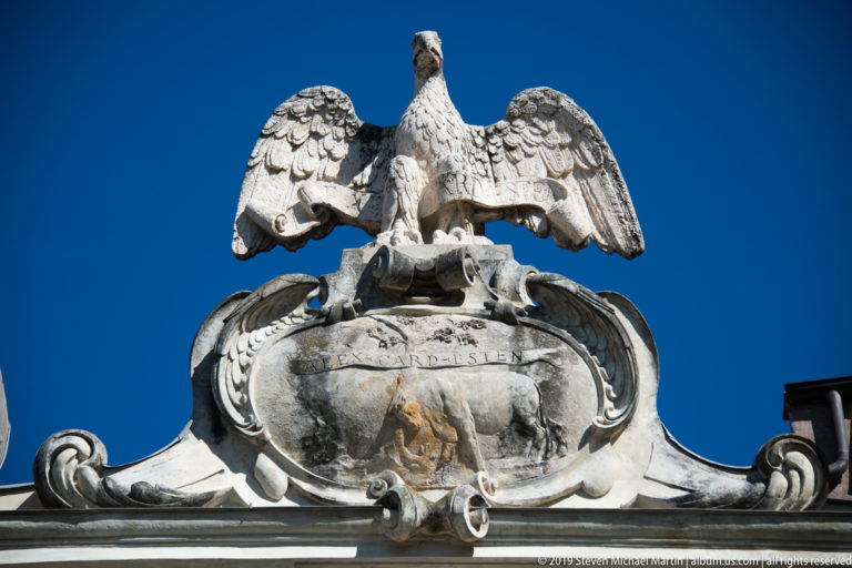 Stone eagle with spread wings perched above a weathered scrolling heraldic cartouche against blue sky.