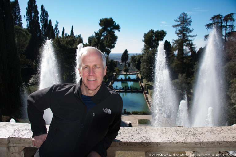 Man in black jacket at a stone parapet with tall fountain jets and rectangular garden pools behind him.