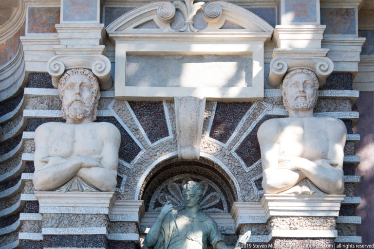 Two carved male support figures flank an arched niche in an ornate striped-stone fountain façade.