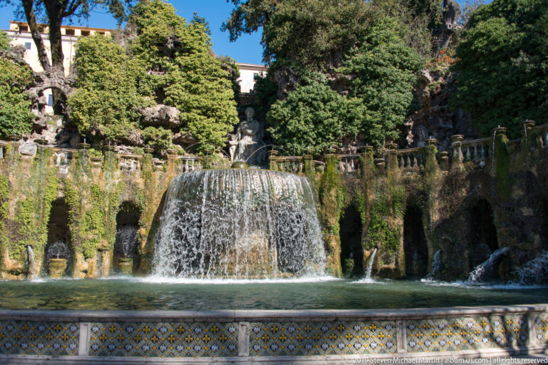 Frontal view across a tiled pool edge toward a broad cascade, grotto wall, balustrade, and trees.