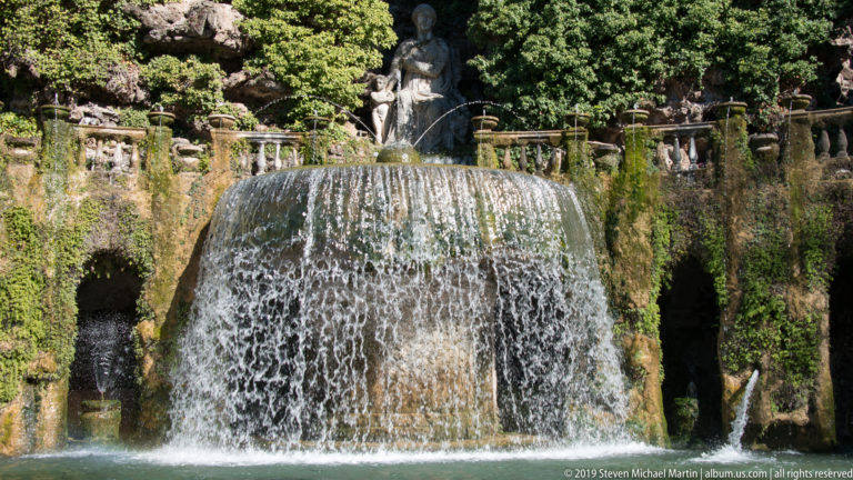 Broad curtain of water falling into a pool beneath an upper balustrade and standing statue.