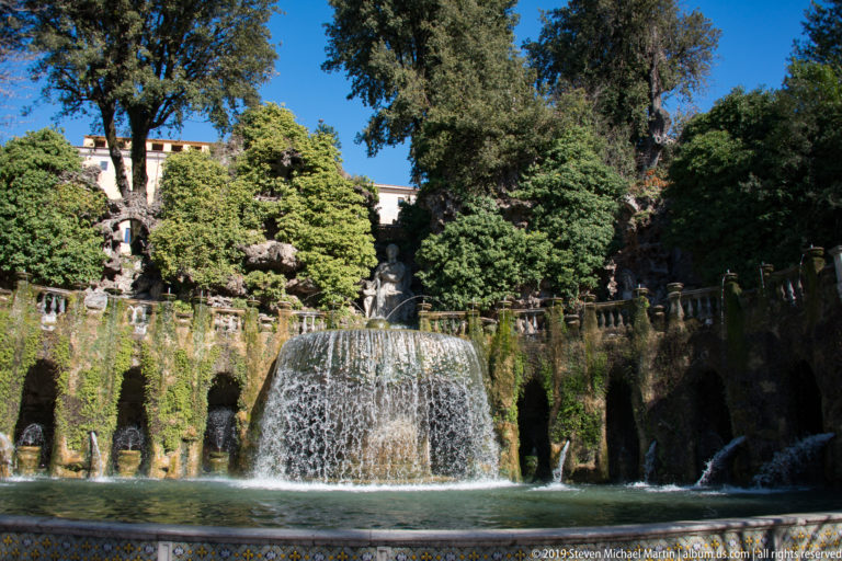 Wide garden view of a broad central cascade, pool, grotto openings, balustrade, and dense trees.
