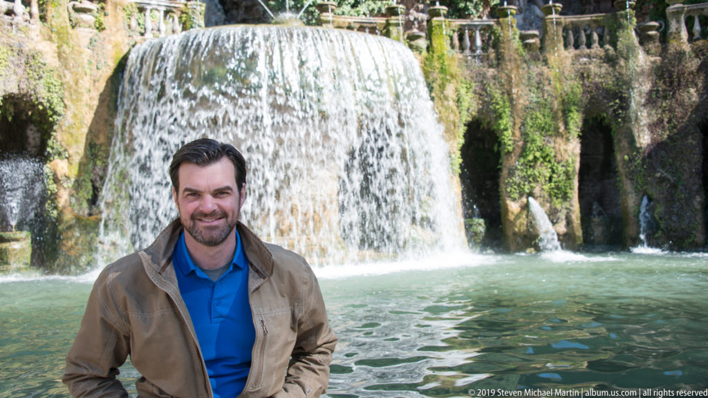 Man in a blue shirt and tan jacket standing before a broad fountain cascade and green pool.