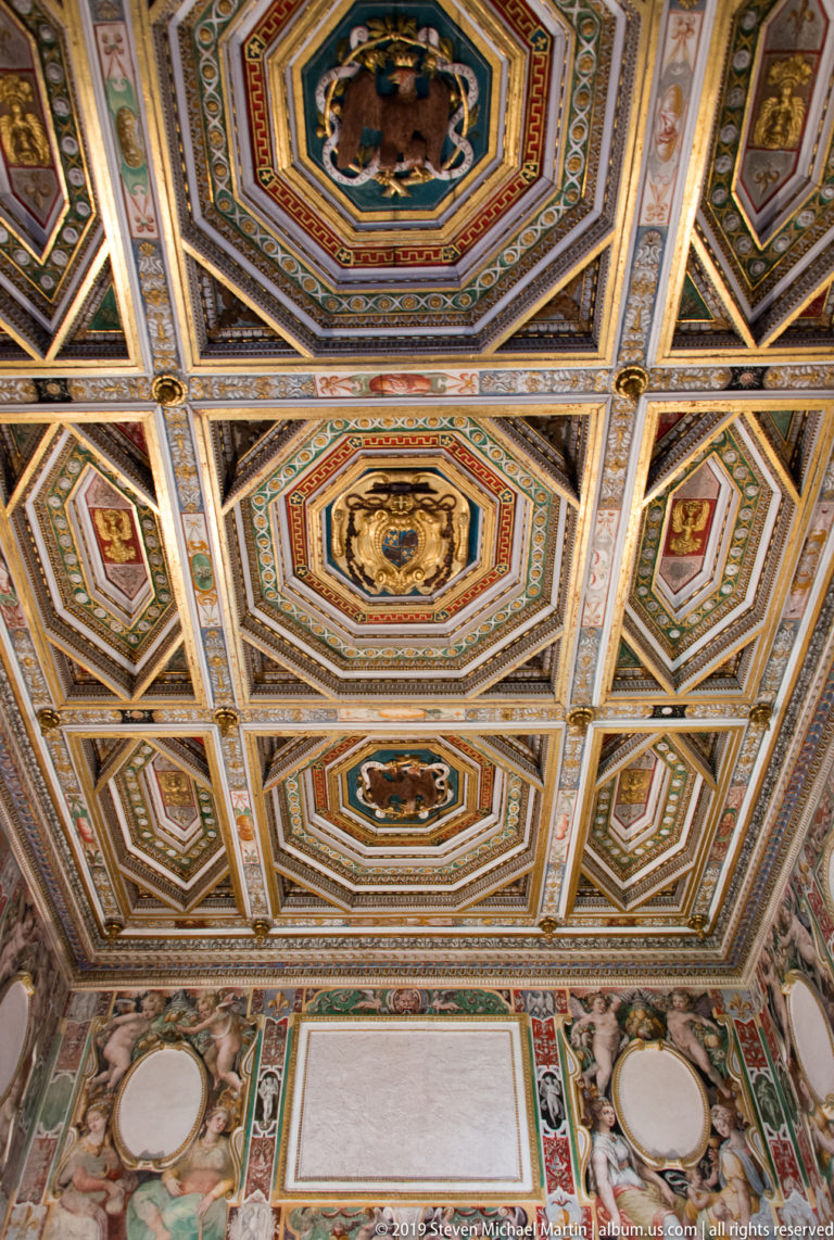 Geometric coffered ceiling of polygonal painted compartments with small figurative medallions and patterned moldings.