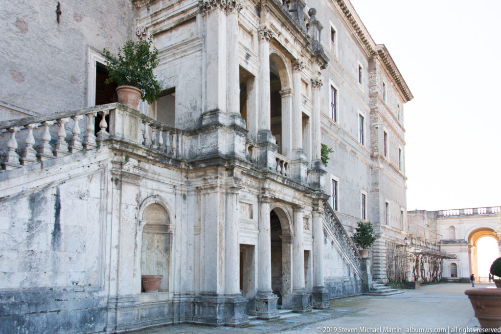 Weathered Renaissance palace façade with pilasters, arched openings, balustrade, stair, and sunlit passage at right.