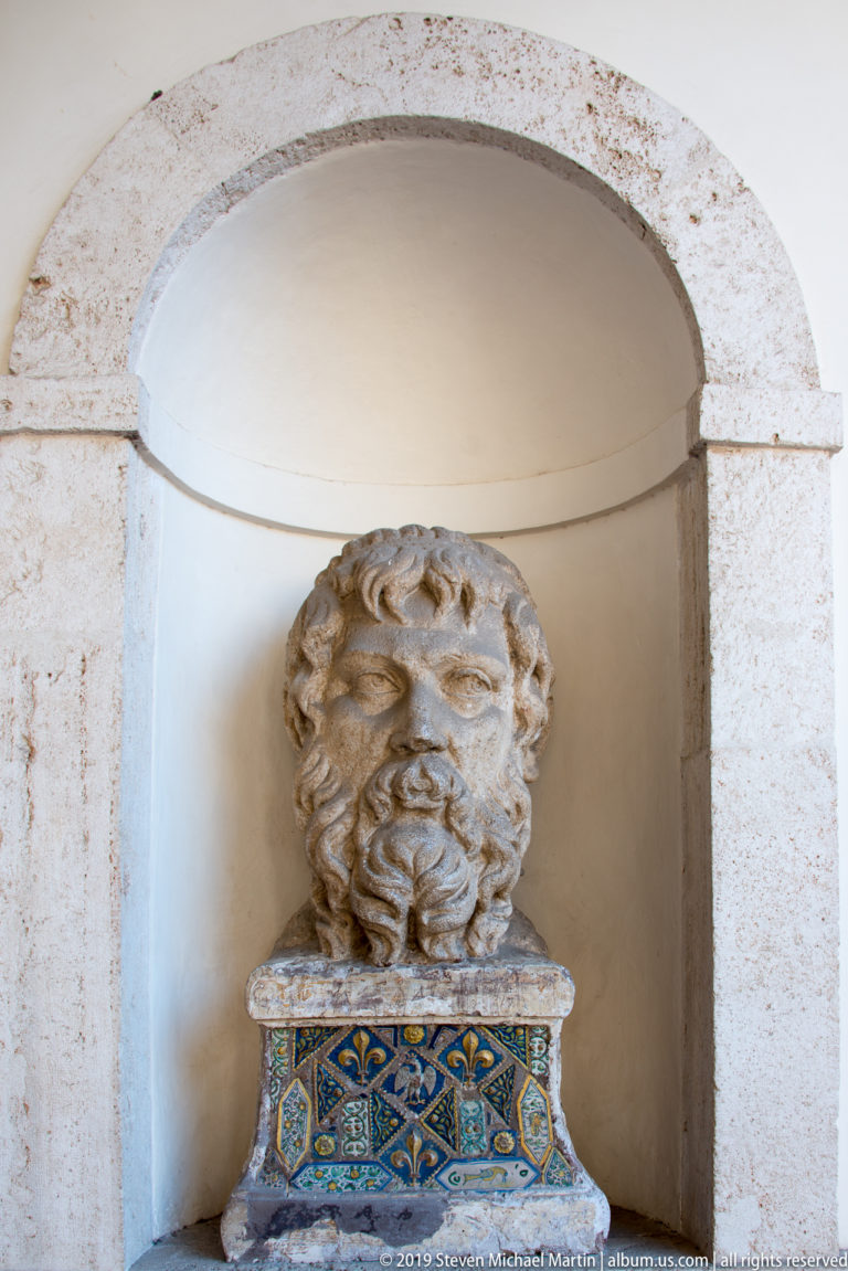 Weathered bearded stone head in an arched niche above a blue-and-yellow patterned ceramic base.