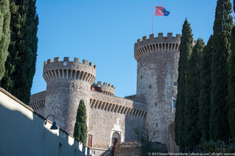 Stone fortress with two round crenellated towers, cypress trees, and an ascending approach to an arched entrance.