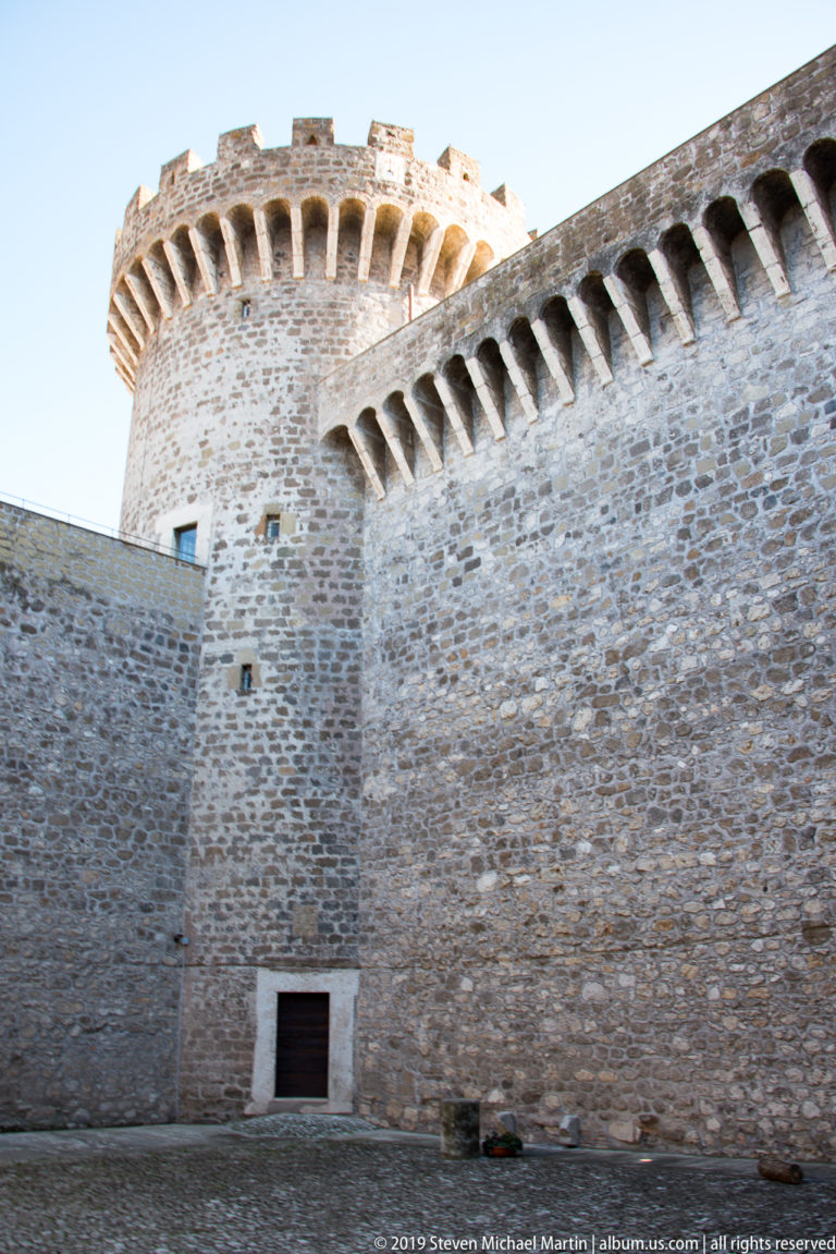 Rough stone fortress wall and round crenellated tower rising above an empty interior courtyard.