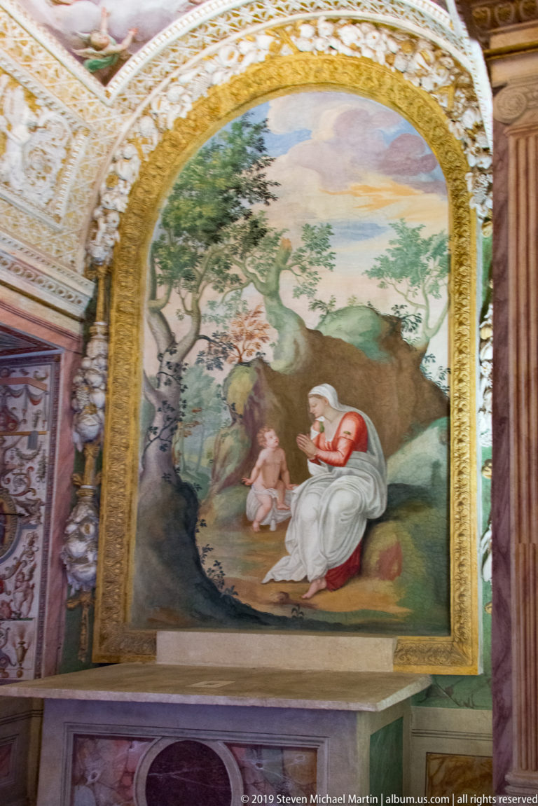 Pastoral fresco of a seated woman in pale robes with a child beneath a large tree and distant hills.