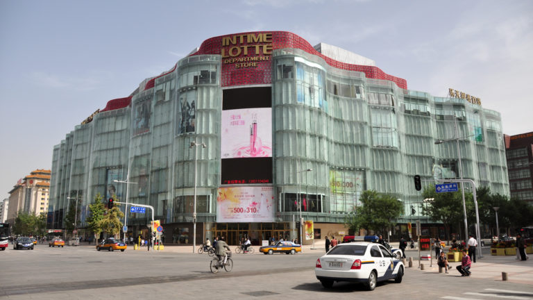 Curved glass exterior of the Intime Lotte department store at a busy intersection with cars, bicycles, and pedestrians in Beijing.