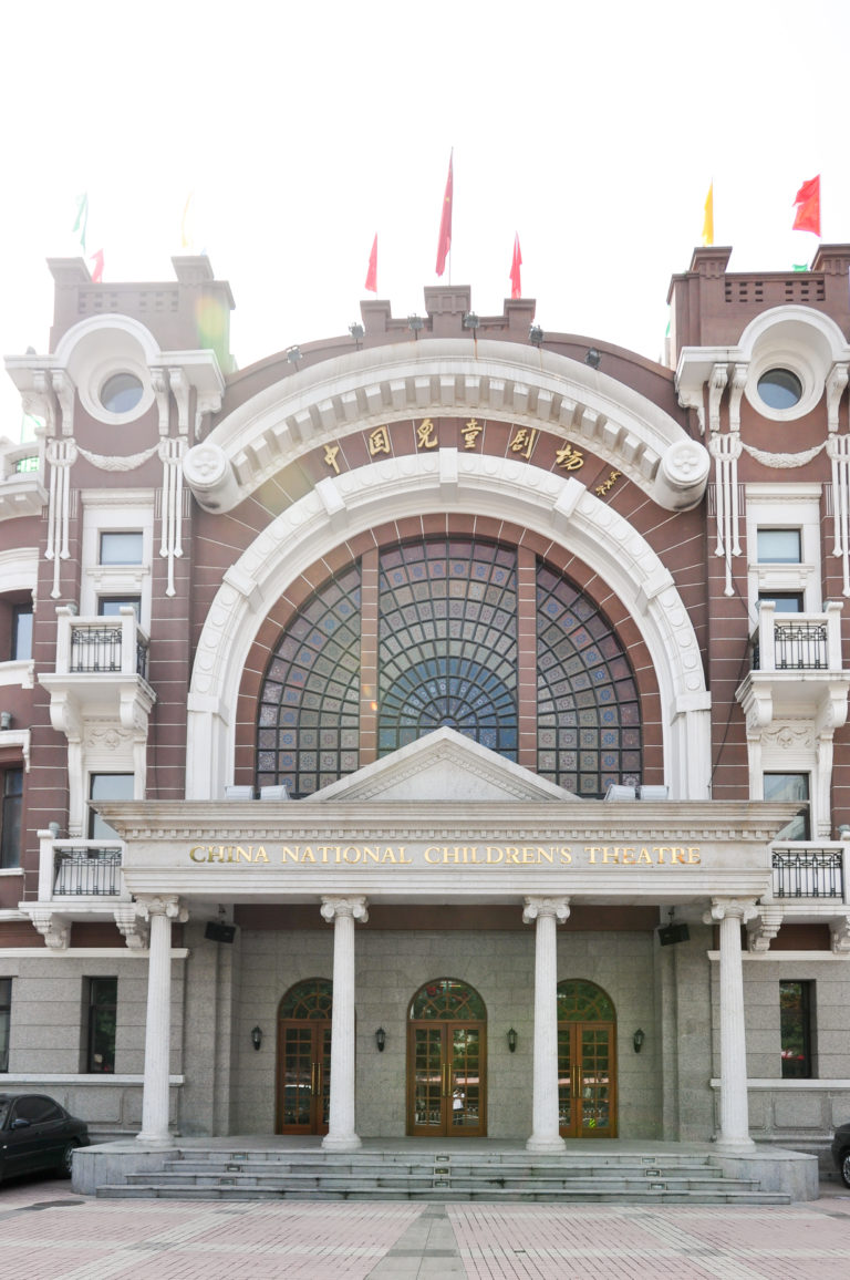 Front façade of the China National Children's Theatre in Beijing, with a large arched window, columns, balconies, entrance doors, and rooftop flags.
