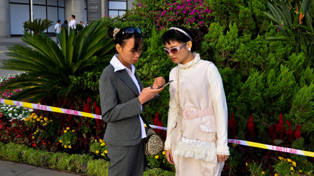 Two women stand beside colorful flower beds and dense ornamental greenery in Beijing, one looking down at an object in her hands.