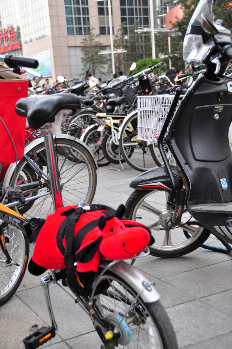 Parked bicycles and motor scooters on a Beijing sidewalk, including a bicycle with a bright red-and-black ladybug-shaped accessory.