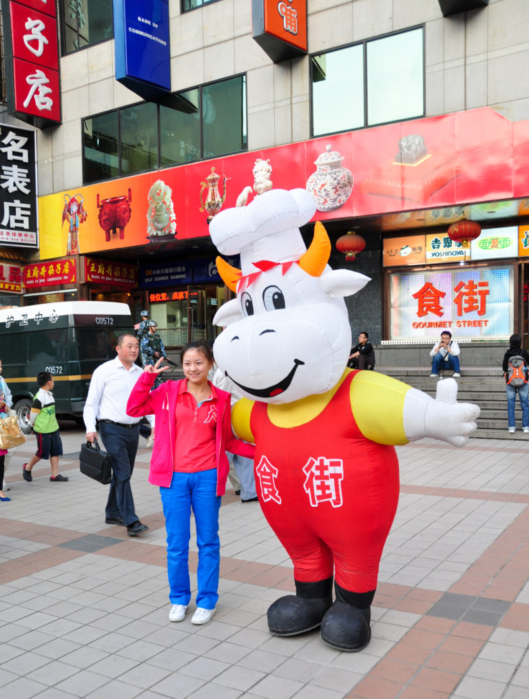 Woman posing beside a large white cow mascot in a chef’s hat and red 食街 shirt outside a building marked Gourmet Street.