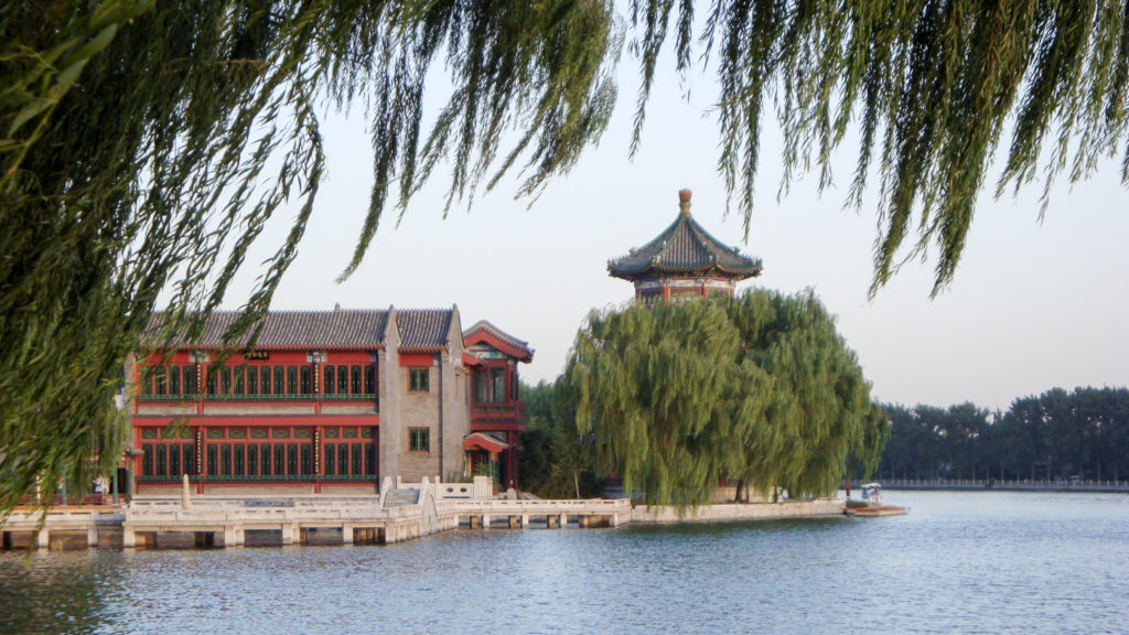 Traditional multi-story building and pavilion among willow trees beside a calm lake in Beijing.