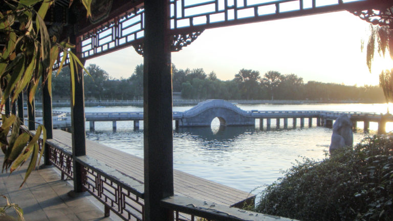 Pale arched moon bridge over a calm lake, seen through the posts of a lakeside pavilion, with the bridge’s arch reflected to form a circular motif.