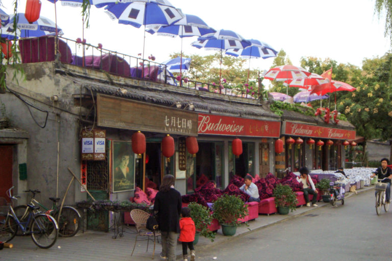 Canal-side Houhai bar in Beijing with Budweiser signs, red lanterns, flowers, and a rooftop terrace with umbrellas.