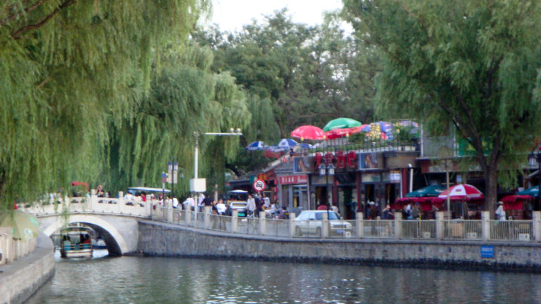 Lakeside cafes with colorful umbrellas beside a low stone bridge along the Shichahai waterfront in Beijing’s Houhai district.