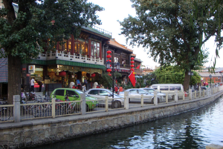 Traditional-style restaurant with red lanterns beside a canal road with cars and bicycles in the Shichahai area of Beijing.