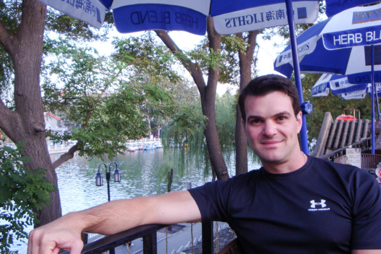Dark-haired traveler seated and smiling at a lakeside café in Beijing, with water and willow trees behind him.
