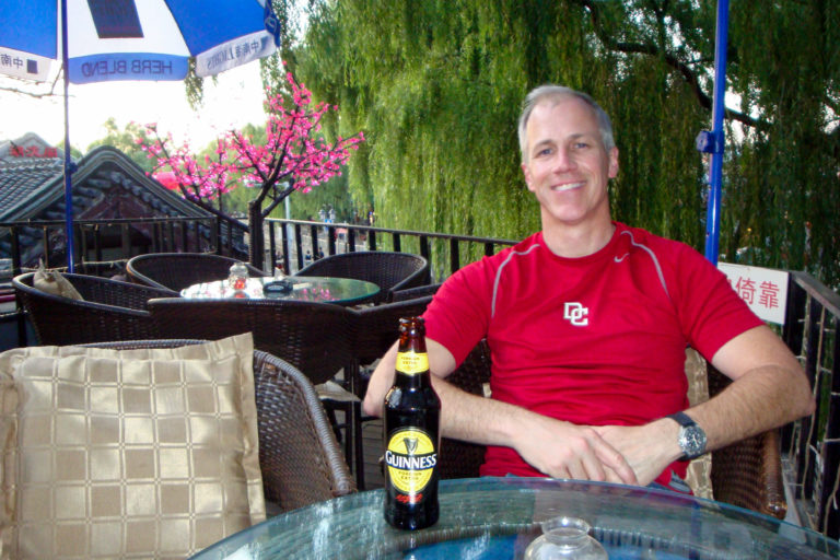 Gray-haired traveler in a red shirt seated at an outdoor lakeside cafe table with a Guinness bottle.