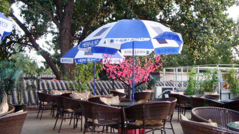 Wicker tables and chairs beneath blue-and-white HERB BLEND umbrellas on a lakeside terrace by a Beijing lake.