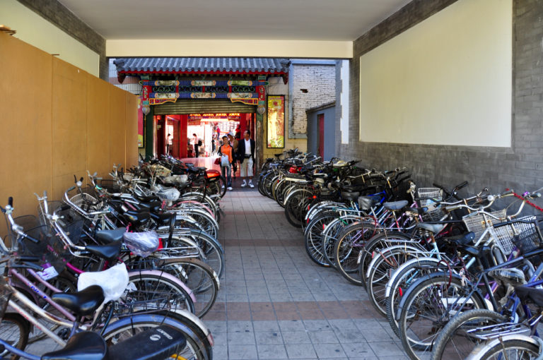 Rows of bicycles parked wheel-to-wheel in a covered passage beneath a painted traditional gateway in Beijing.