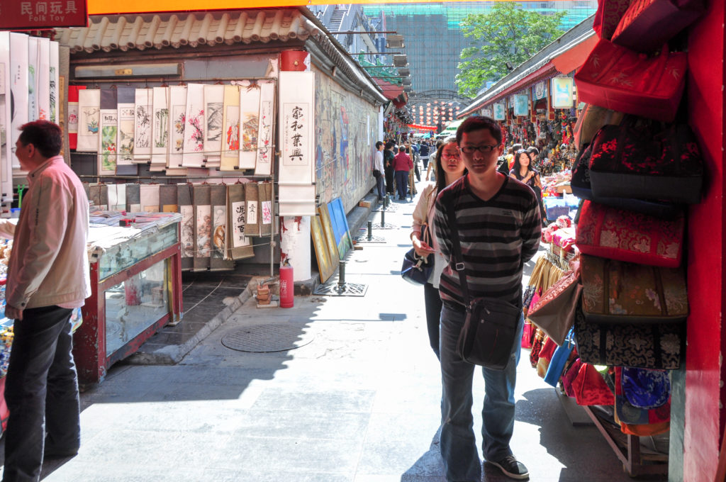 Man walking toward the camera through a narrow Beijing market passage lined with paintings and hanging bags.