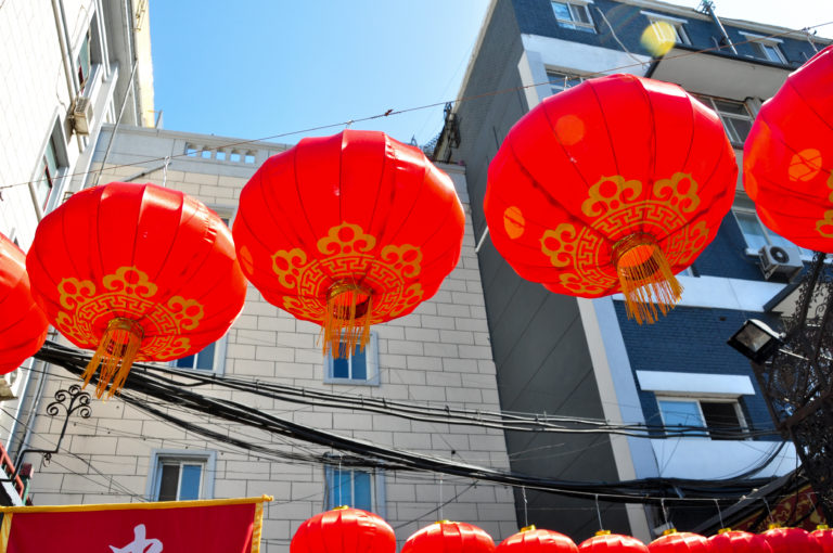 Oversized red lanterns with gold patterns and tassels hanging above a Beijing market street.