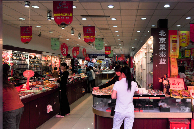 Indoor Beijing jewelry and handicraft market with shoppers examining decorative objects and cloisonné-related goods in glass counters under bright lights.
