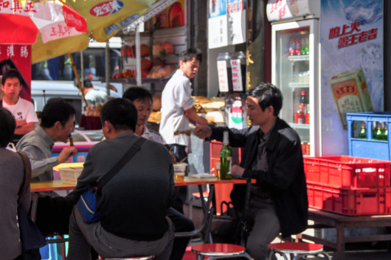 Men eating at bright red outdoor tables on low stools beside a neighborhood restaurant in Beijing.