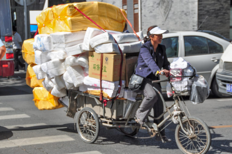 Woman riding a heavily loaded three-wheeled cargo tricycle through city traffic, with white and yellow packages stacked high on the rear platform.