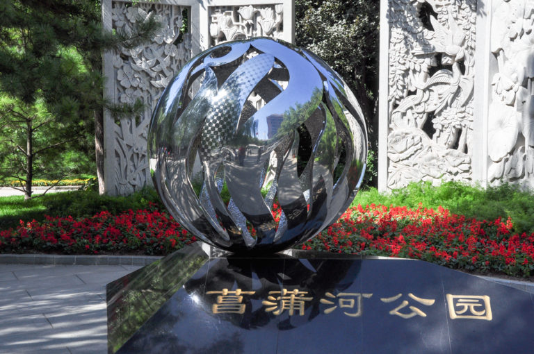 Reflective metal sphere sculpture above a black stone sign reading 青年湖公园 at the entrance to Qingnianhu Park, with carved stone relief panels in the background.